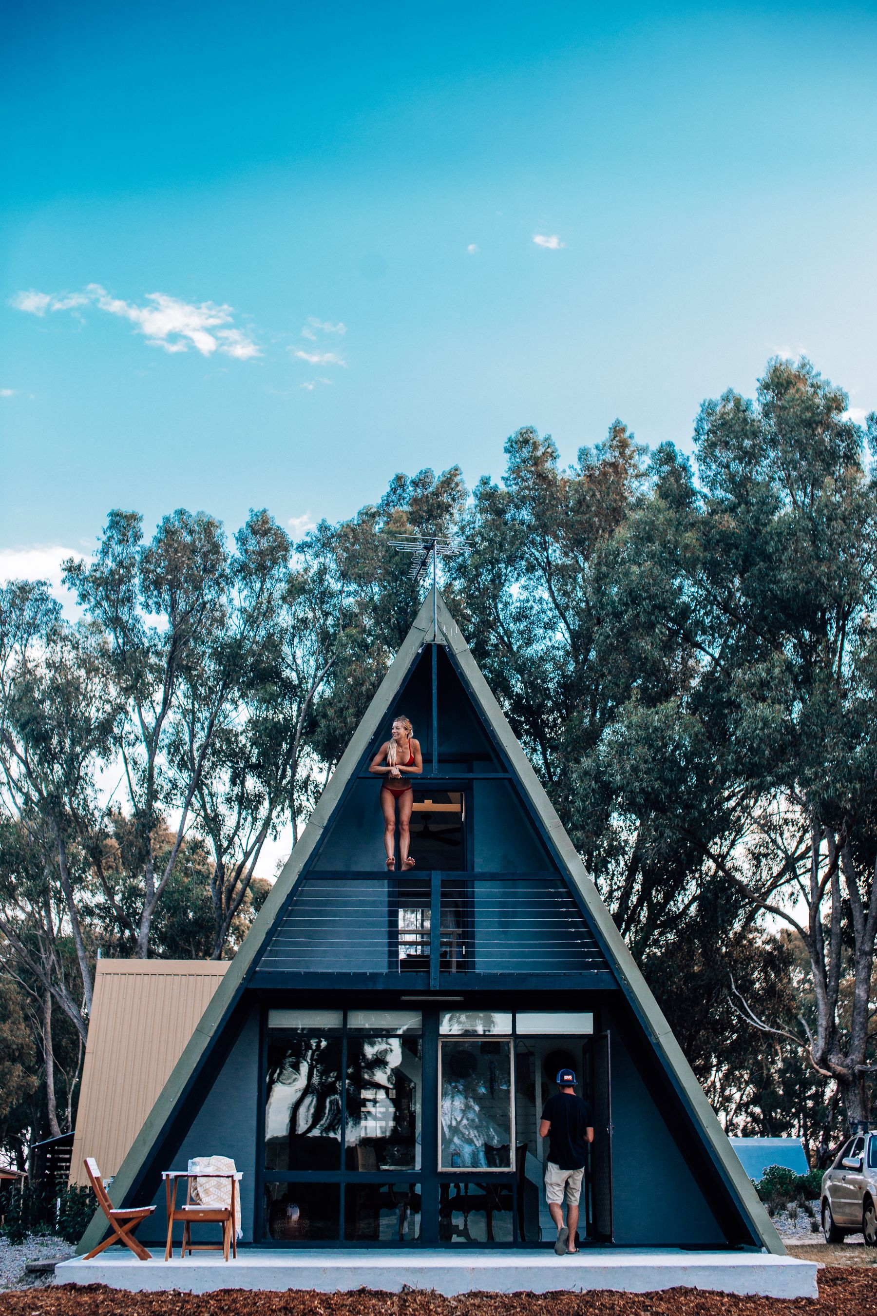 Person sitting outside a blue A-frame vacation rental cabin surrounded by trees